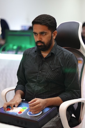 A person is seated in a chair, focused intently on something, with hands positioned on a gaming console or arcade controller. The setup appears to be indoors, with visible screens and lighting in the background, suggesting an electronic gaming environment.