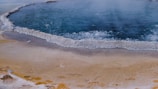 A geothermal pool surrounded by a white mineral crust. The water is dark blue and appears to be bubbling, indicating high temperatures. The surrounding ground is a mix of beige and yellowish hues with some wet patches.