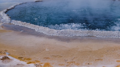A geothermal pool surrounded by a white mineral crust. The water is dark blue and appears to be bubbling, indicating high temperatures. The surrounding ground is a mix of beige and yellowish hues with some wet patches.