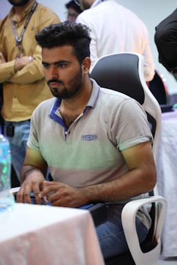 A man with a focused expression is seated in a black and white ergonomic chair. He wears a light gray polo shirt with a logo and operates a keyboard. The background shows several people standing, partially visible, suggesting a social or competitive setting.
