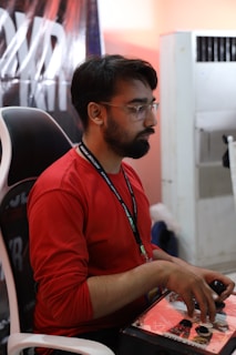 A person with glasses, wearing a red shirt and a lanyard, is sitting on a chair and focused on playing a video game using an arcade joystick setup. The environment includes a partially visible air conditioner and a poster in the background.