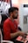 A person with glasses, wearing a red shirt and a lanyard, is sitting on a chair and focused on playing a video game using an arcade joystick setup. The environment includes a partially visible air conditioner and a poster in the background.