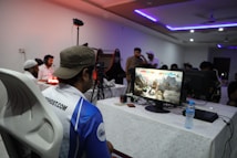 A group of people is gathered in a room watching and participating in a video game competition. Players and spectators are focused on the monitors displaying a fighting game. The setting includes tables with equipment and chairs arranged around the venue, with a noticeable intensity in the participants’ engagement.