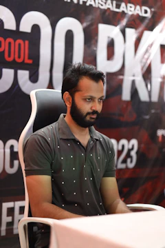 A man with a beard and mustache is seated on a white chair with a black backrest. He is wearing a gray polo shirt with small white dots. The background shows a large, partially visible banner with text that includes 'POOL' and 'PK'.