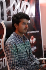 A person is sitting on a gaming chair wearing a checkered shirt. Behind him, a large poster with partially visible letters and logos is displayed.