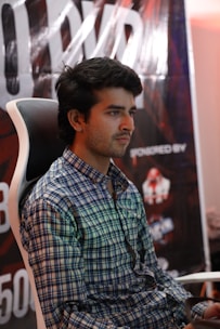 A person is sitting on a gaming chair wearing a checkered shirt. Behind him, a large poster with partially visible letters and logos is displayed.