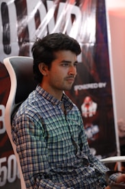 A person is sitting on a gaming chair wearing a checkered shirt. Behind him, a large poster with partially visible letters and logos is displayed.