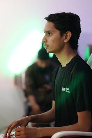 A person is sitting in a focused posture, possibly engaging with a gaming console. The lighting is dim with a green glow, creating a calm yet intense atmosphere. Another person is blurred in the background.