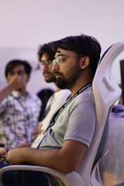 Three people are seated indoors, possibly in a conference or meeting setting. The focus is on a person wearing glasses and a lanyard, sitting in a chair with a serious expression.