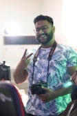 A person with glasses and a beard is smiling and holding a Canon camera. They are wearing a patterned shirt with blue leaf designs. The person is making a peace sign with one hand. The setting appears to be indoors with soft lighting, and there is a hint of green light reflecting on the person.