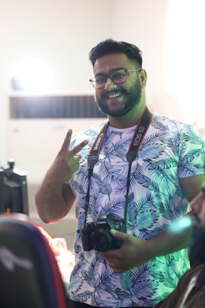 A person with glasses and a beard is smiling and holding a Canon camera. They are wearing a patterned shirt with blue leaf designs. The person is making a peace sign with one hand. The setting appears to be indoors with soft lighting, and there is a hint of green light reflecting on the person.