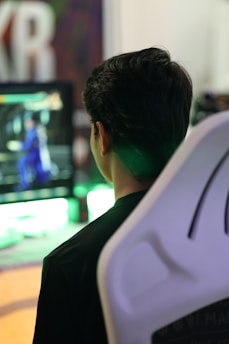 A person is sitting in a white gaming chair while looking at a screen displaying a video game with a character in blue. The background includes blurred colors and large, out-of-focus lettering.