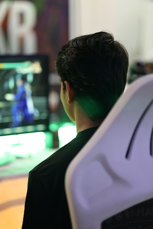 A person is sitting in a white gaming chair while looking at a screen displaying a video game with a character in blue. The background includes blurred colors and large, out-of-focus lettering.