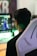 A person is sitting in a white gaming chair while looking at a screen displaying a video game with a character in blue. The background includes blurred colors and large, out-of-focus lettering.