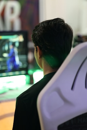 A person is sitting in a white gaming chair while looking at a screen displaying a video game with a character in blue. The background includes blurred colors and large, out-of-focus lettering.