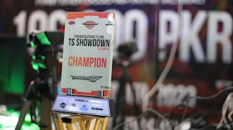 A trophy with the text 'Congratulations to our TS Showdown Season 04 Champion' and 'Tekken 7' is prominently displayed. It appears to be an award for a gaming competition that took place in October 2023. In the background, there are blurred images of gaming equipment and signs with large texts.
