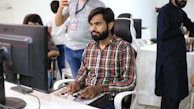 A man with a beard, wearing a plaid shirt, is seated at a desk, focused on a gaming controller or similar device. He is surrounded by other people in a casual office setting, with a large computer monitor in front of him. The background shows a mix of people standing and sitting, engaged in various activities.
