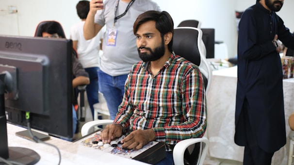 A man with a beard, wearing a plaid shirt, is seated at a desk, focused on a gaming controller or similar device. He is surrounded by other people in a casual office setting, with a large computer monitor in front of him. The background shows a mix of people standing and sitting, engaged in various activities.