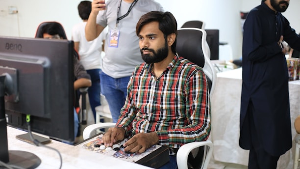 A man with a beard, wearing a plaid shirt, is seated at a desk, focused on a gaming controller or similar device. He is surrounded by other people in a casual office setting, with a large computer monitor in front of him. The background shows a mix of people standing and sitting, engaged in various activities.