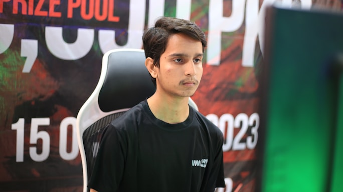 A young man in a black shirt sits in front of a computer monitor, deeply focused. Behind him, a colorful background with text hints at a competitive or event setting.