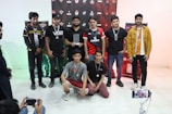 A group of eight young men are gathered indoors, some standing and some kneeling in front. They are casually dressed, wearing medals around their necks and one holds a trophy. Behind them, a decorative backdrop featuring logos and graphics is visible. Two individuals on the sides appear to be taking photos or videos with smartphones.