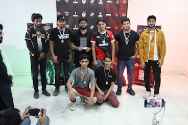 A group of eight young men are gathered indoors, some standing and some kneeling in front. They are casually dressed, wearing medals around their necks and one holds a trophy. Behind them, a decorative backdrop featuring logos and graphics is visible. Two individuals on the sides appear to be taking photos or videos with smartphones.