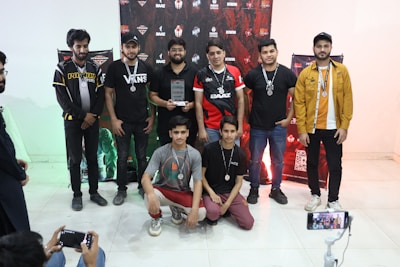 A group of eight young men are gathered indoors, some standing and some kneeling in front. They are casually dressed, wearing medals around their necks and one holds a trophy. Behind them, a decorative backdrop featuring logos and graphics is visible. Two individuals on the sides appear to be taking photos or videos with smartphones.
