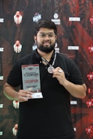 A man with glasses and a beard is holding a trophy and a medal. He is wearing a black shirt and a lanyard. The background is a branded backdrop with various logos and texts, predominantly in red and black colors.