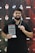 A man with glasses and a beard is holding a trophy and a medal. He is wearing a black shirt and a lanyard. The background is a branded backdrop with various logos and texts, predominantly in red and black colors.