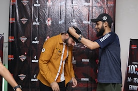 Two people are engaged in a light-hearted interaction where one is placing a cap on the other's head. The background features a wall adorned with logos and branding related to an event or competition.