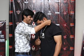 Two people are participating in an award ceremony. One person is placing a medal around the neck of another, suggesting a celebratory or recognition event. Both individuals appear to be in good spirits, and the background features branded banners and logos associated with a particular event or organization.