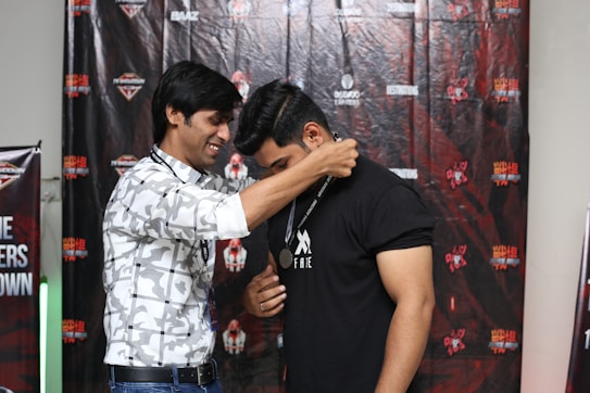 Two people are participating in an award ceremony. One person is placing a medal around the neck of another, suggesting a celebratory or recognition event. Both individuals appear to be in good spirits, and the background features branded banners and logos associated with a particular event or organization.