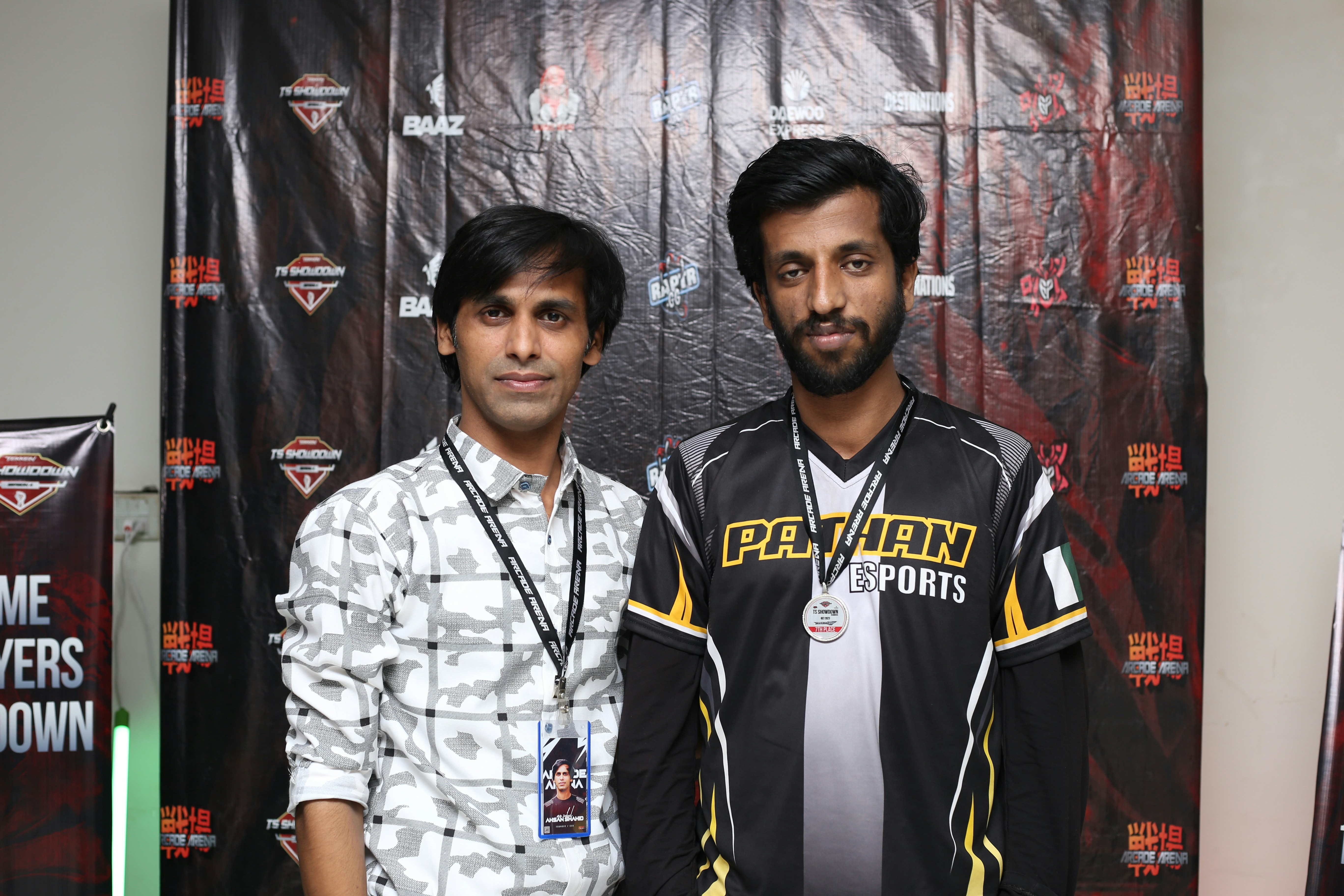 Two individuals stand in front of a backdrop with logos and text, dressed in casual clothing and sporting lanyards with badges. The individual on the left wears a patterned shirt, while the one on the right wears a shirt with the words 'Pathan Esports'.