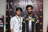 Two individuals stand in front of a backdrop with logos and text, dressed in casual clothing and sporting lanyards with badges. The individual on the left wears a patterned shirt, while the one on the right wears a shirt with the words 'Pathan Esports'.