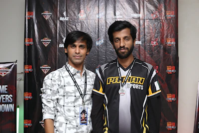 Two individuals stand in front of a backdrop with logos and text, dressed in casual clothing and sporting lanyards with badges. The individual on the left wears a patterned shirt, while the one on the right wears a shirt with the words 'Pathan Esports'.