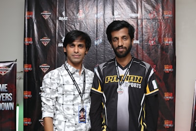Two individuals stand in front of a backdrop with logos and text, dressed in casual clothing and sporting lanyards with badges. The individual on the left wears a patterned shirt, while the one on the right wears a shirt with the words 'Pathan Esports'.