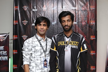 Two individuals stand in front of a backdrop with logos and text, dressed in casual clothing and sporting lanyards with badges. The individual on the left wears a patterned shirt, while the one on the right wears a shirt with the words 'Pathan Esports'.