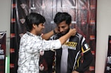 Two men are standing in front of a backdrop featuring various logos and text. One man is placing a medal or lanyard around the neck of the other man, who is wearing a black and yellow sports jersey with the word 'KHAN' visible.