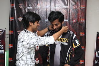 Two men are standing in front of a backdrop featuring various logos and text. One man is placing a medal or lanyard around the neck of the other man, who is wearing a black and yellow sports jersey with the word 'KHAN' visible.