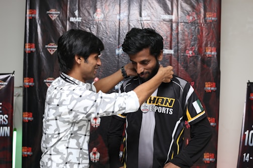 Two men are standing in front of a backdrop featuring various logos and text. One man is placing a medal or lanyard around the neck of the other man, who is wearing a black and yellow sports jersey with the word 'KHAN' visible.