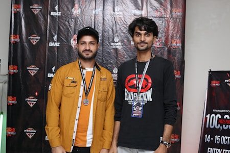 Two men stand side by side in front of a backdrop with various logos and text. One man is wearing a mustard yellow jacket over a white and orange outfit, with a medal around his neck. The other man is wearing a black sweatshirt with a red and black graphic, and both have lanyards around their necks. The backdrop includes branding and promotional content.