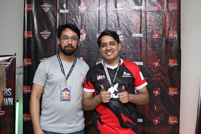 Two men are standing together in front of a backdrop with various logos and text. The man on the left is wearing a light gray shirt and glasses, holding an ID badge on a lanyard. The man on the right is smiling and wearing a red and black jersey, holding up a medal around his neck.