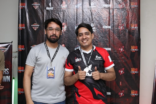 Two men are standing together in front of a backdrop with various logos and text. The man on the left is wearing a light gray shirt and glasses, holding an ID badge on a lanyard. The man on the right is smiling and wearing a red and black jersey, holding up a medal around his neck.