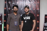 Two men are standing in front of a backdrop decorated with logos and text. One man is wearing a striped shirt with glasses and a badge, and the other is wearing a black t-shirt with the word 'VANS' and a cap. Both have lanyards around their necks.