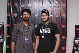 Two men are standing in front of a backdrop decorated with logos and text. One man is wearing a striped shirt with glasses and a badge, and the other is wearing a black t-shirt with the word 'VANS' and a cap. Both have lanyards around their necks.