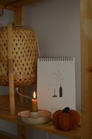 A wooden shelf holds a wicker lamp, a 2024 calendar with minimalist designs, a lit candle in a ceramic holder, and a knitted pumpkin decoration. The arrangement creates a warm and cozy atmosphere.