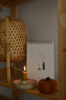 A wooden shelf holds a wicker lamp, a 2024 calendar with minimalist designs, a lit candle in a ceramic holder, and a knitted pumpkin decoration. The arrangement creates a warm and cozy atmosphere.