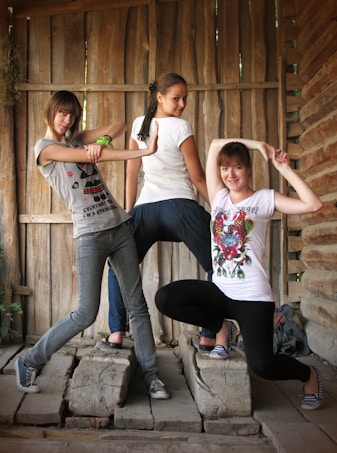 Three young women are striking playful poses inside a rustic wooden structure. They are standing on uneven wooden beams, and their expressions and poses suggest a sense of fun and camaraderie. The setting appears natural with wooden walls forming the backdrop.
