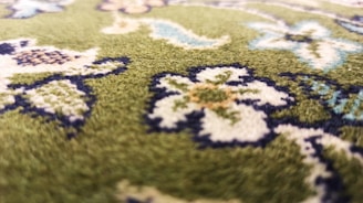 A close-up view of a green carpet with intricate floral patterns. The design includes flowers and leaves in shades of white, blue, and brown, outlined in dark blue. The texture of the carpet is visible, suggesting a soft, plush surface.