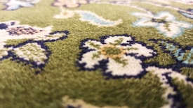 A close-up view of a green carpet with intricate floral patterns. The design includes flowers and leaves in shades of white, blue, and brown, outlined in dark blue. The texture of the carpet is visible, suggesting a soft, plush surface.